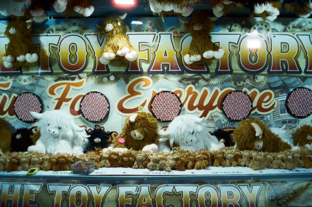 A colorful toy booth named 'The Toy Factory' featuring various plush animals, including white and brown fluffy toys, displayed on a table amidst decorative gears and patterns.